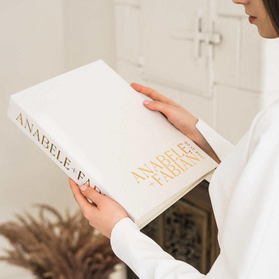 A person holding a white and gold wedding guest book with the name 'Anabele + Fabian' printed on the cover.