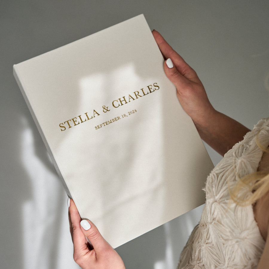 A person holding a white wedding guest book with gold text, titled 'STELLA & CHARLES' on the cover.